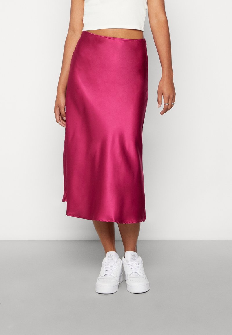 Femme portant une jupe midi en satin rose vif, un crop top blanc et des baskets blanches, debout devant un fond clair uni.