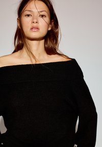 Zwarte off-shoulder trui met een geribde halslijn en een ontspannen pasvorm. De textuur lijkt zacht en geschikt voor casual gebruik.