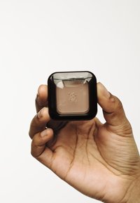 Vierkante, zwarte compact in een hand, met daarin een lichtbruine poeder, met een doorzichtige deksel en een gegraveerd logo op het productoppervlak.