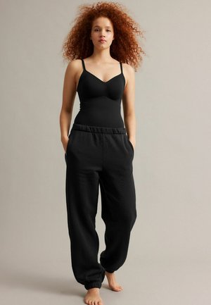 Ung kvinde med krøllet rødt hår iført en sort camisole og løse sorte joggingbukser, stående barfodet mod en ensfarvet baggrund.