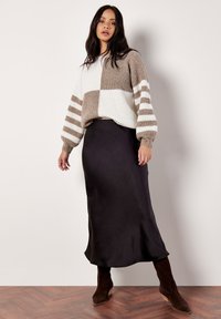 Sweter dzianinowy w beżowo-białym kolorze z wzorem w bloki i paskowanymi rękawami, zestawiony z elegancką czarną satynową spódnicą oraz brązowymi botkami na obcasie.