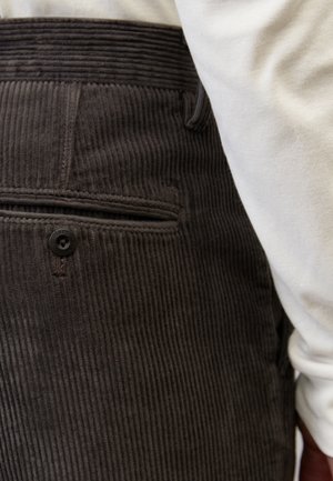Tummanruskeat corduroy-housut, joissa pystysuora ribbineulos; niissä on takatasku napillisella suljennalla ja korkeavyötäröinen vyötärönauha. Valkoinen paita osittain näkyvissä.