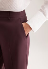 Bordeauxkleurige broeken met een slanke pasvorm, voorzien van een zijzak en een gladde textuur. Gecombineerd met een witte, gestreepte langemouwen shirt.