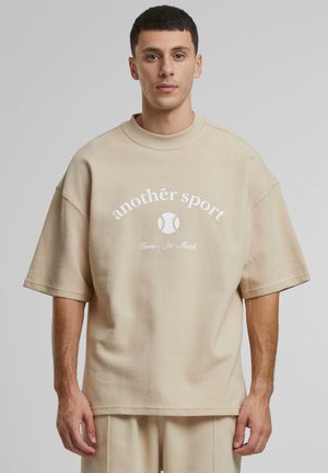 Beige oversized tekstureret t-shirt med rund hals. Har hvidt trykt tekst og grafik af en tennisbold. Kortærmet og afslappet pasform.