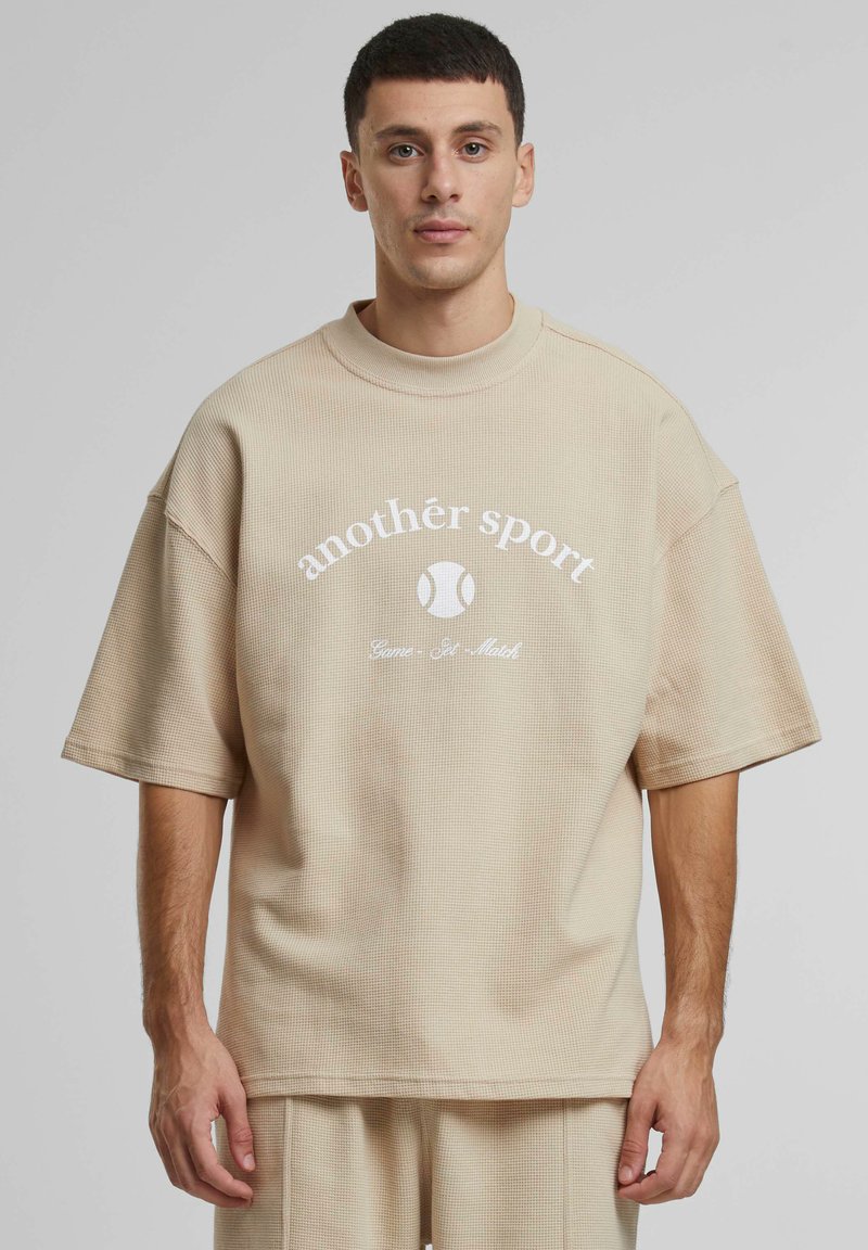 Beige spalvota oversized tekstūruota marškinė su apvalia apykakle. Yra baltas spausdintas tekstas ir teniso kamuoliuko grafika. Trumpomis rankovėmis ir laisva forma.