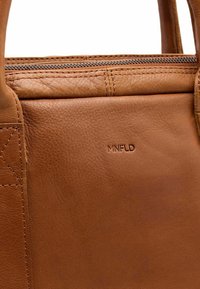 Hellbraune Lederhandtasche mit glatter Oberfläche, ausgestattet mit einem oberen Reißverschluss und geprägtem "MNFLD"-Logo auf der Vorderseite. Schlichtes, minimalistisches Design.
