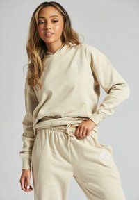 Kobieta z długimi włosami, ubrana w beżowy hoodie i dopasowane dresy z białym logo góry, stojąca na tle o jednolitej fakturze.