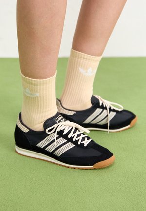 Persona que lleva zapatillas negras Adidas con rayas blancas y calcetines beige con un logo blanco, de pie sobre una alfombra verde.