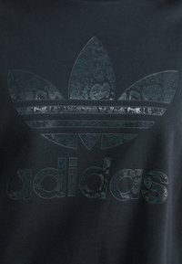 Fekete sportöltözet, amelyen egy textúrázott, fényes fekete kígyóbőr mintázatú logó és szöveg található. A dizájn tartalmaz három levélformájú csíkot a márkaneve felett.