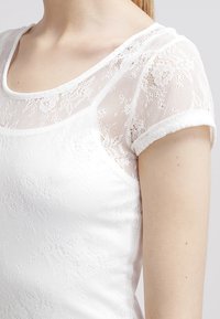 Bílý top s průhlednými krajkovými krátkými rukávy, kulatým výstřihem a přiléhavým trupem. Tkanina má květinový vzor a texturované detaily.