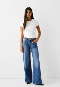 Valkoinen lyhythihainen crop-top, joka on yhdistetty korkeavyötäröisiin, leveälahkeisiin sinisiin farkkuihin. Farkuissa on lievää kulumaa ja perinteiset taskut.