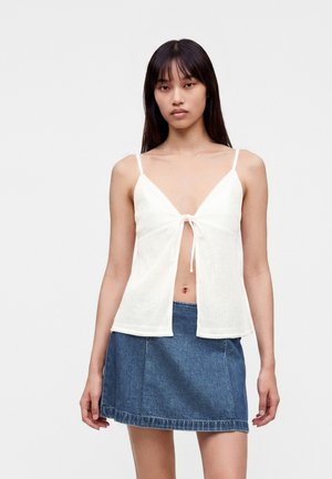 Ung kvinde iført en hvid ærmeløs top med bindebånd foran og en blå denim mininederdel, stående mod en ensfarvet lys baggrund.