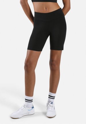 Person trägt schwarze, hoch taillierte Radlerhosen, weiße Sportsocken mit schwarzen Streifen und weiße Sneaker und steht vor einem schlichten Hintergrund.