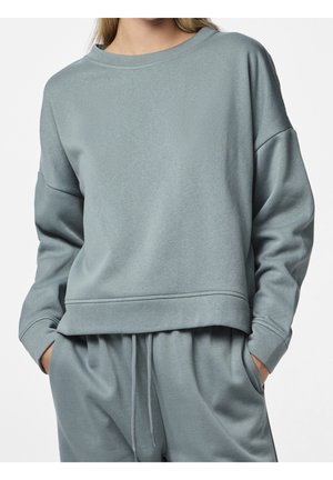 Frau trägt ein hellgraues, übergroßes Sweatshirt und passende Jogginghosen mit Kordelzug, die Hände in den Taschen.