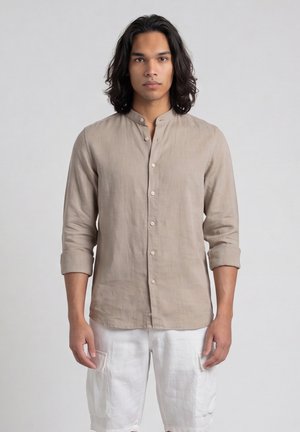 Jonge man met schouderlang donker haar, gekleed in een beige overhemd met knopen en opgerolde mouwen en witte cargoshorts, staand tegen een effen achtergrond.