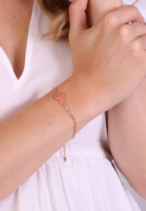 Gouden armband met een roze hartvormige edelsteen, uitgerust met een delicate schakeldesign en een verstelbare sluiting. Draagt om een lichte huidtonen pols.