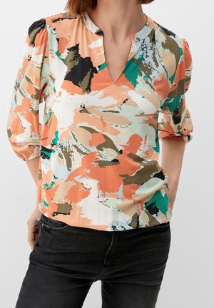 Femme portant un chemisier à col en V avec un motif abstrait orange, vert, beige et noir, manches trois-quarts bouffantes, associé à un jean noir.