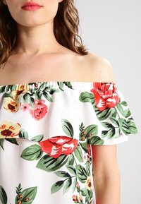 Olkaimeton toppi valkoisesta kankaasta, jossa on kukkakuvio punaisilla ja keltaisilla kukilla sekä vihreillä lehdillä; röyhelökaulus.