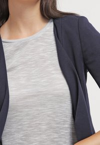 Marineblauer Blazer über einem hellgrauen strukturierten Oberteil. Der Blazer hat ein tailliertes Design mit offenem Vorderteil und weichem Stoff.