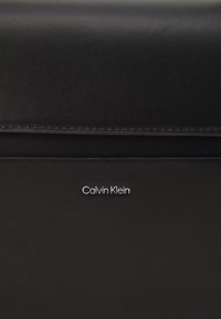 Bolsa de couro preto com uma textura suave e um proeminente logo prateado "Calvin Klein". O design destaca linhas limpas e um estilo minimalista.