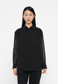 Zwarte blouse met transparante mouwen, knoopsluiting aan de voorkant en spitse kraag. Gladde textuur met een ontspannen pasvorm en afgeronde zoom.