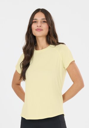 Woman with long dark hair wearing a light yellow short-sleeve shirt and black pants, standing with hands behind her back.
