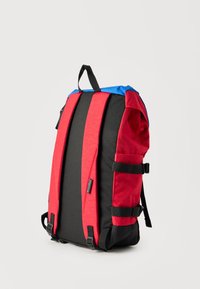 Πίσω όψη ενός κόκκινου και μαύρου σακιδίου με μπλε επάνω μέρος, μαξιλαρωμένους ιμάντες ώμων, πλευρικές αγκράφες και χειρολαβή στην κορυφή.