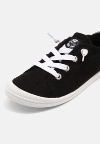 Baskets en toile noire avec semelle en caoutchouc blanche, lacets blancs et patch logo sur la languette, sur un fond clair.