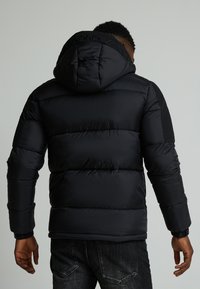 Schwarze Pufferjacke mit Kapuze; gestepptes Design, gerippte Bündchen und Logodetail an der Kapuze. Glatte Textur, mittellange Passform.
