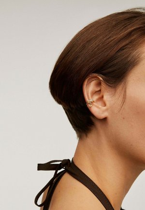 Pendientes de oro con un diseño en espiral, enmarcados por un cabello corto y oscuro. El accesorio está sujeto a la oreja, con un brillo metálico visible.