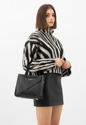 Vrouw met een zwart-wit gestreepte trui en een zwarte leren rok, die een zwarte leren handtas vasthoudt, tegen een witte achtergrond.