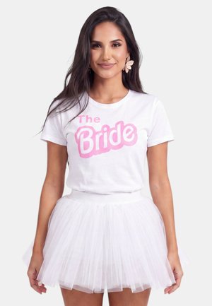 Mujer con cabello largo y oscuro lleva una camiseta blanca con texto rosa "The Bride" y una falda de tul blanca, sonriendo con pendientes en forma de flor.