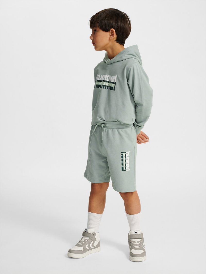 Junge steht mit den Händen hinter dem Rücken, trägt einen hellgrünen Kapuzenpullover und Shorts mit Sportlogo, weiße Socken und graue High-Top-Sneakers.