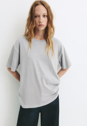 Grå bomullströja med korta ärmar, rund halsringning, avslappnad passform och mjuk textur; kombinerad med mörka jeans.