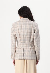 Blazer en tartan beige avec une taille ajustée et des plis dans le dos. Le matériau semble texturé ; possède des accents bleus et bruns, ainsi que des poignets boutonnés.