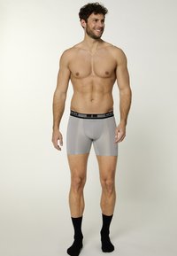 Un uomo sorridente indossa boxer grigi e calze nere, mostrando una posa sicura su uno sfondo chiaro.
