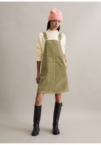 Sukienka na szelkach z zielonym sztruksu z regulowanymi paskami, przednią kieszenią, w komplecie z kremowym swetrem i czarnymi kozakami do kolan oraz różową czapką beanie.