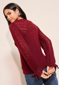 Burgundy stickad tröja med spetsmönstrade ärmar, ribbade manschetter knutna med rosetter och en strukturerad baksida. Avslappnad passform med en mjuk finish.