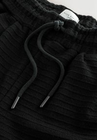 Schwarze Jogginghosen mit strukturiertem Stoff, geripptem Gewebe, verstellbarem Kordelzug mit Metallösen und seitlichen Taschen. Etikett sichtbar im Bund.
