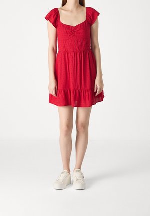 Frau trägt ein rotes kurzärmeliges Kleid mit gerafftem Ausschnitt und weißen Sneakers und steht vor einem schlichten weißen Hintergrund.