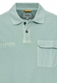 Lichtgroen poloshirt van textuurstof, met een kraag, twee knopen en een zak op de linkerkant van de borst met logo en datumnotatie.