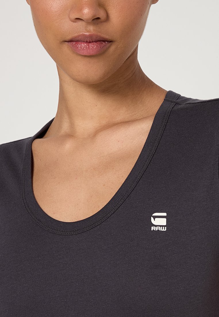 Zbliżenie na osobę w ciemnoszarej koszulce z okrągłym dekoltem, na której piersi widnieje małe białe logo "G RAW".