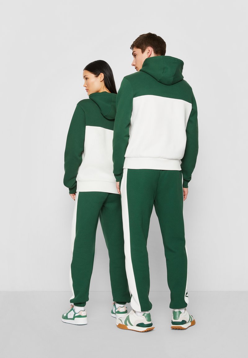 Set di felpa con cappuccio e pantaloni della tuta in cotone verde e bianco, caratterizzato da un design a blocchi di colore, cappuccio, polsini elasticizzati e strisce laterali. Le sneakers sono verdi con accenti bianchi.
