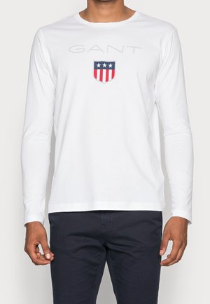Man wearing white long-sleeve shirt with GANT logo and shield emblem with stars and stripes, paired with dark pants.