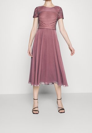 Vrouw draagt een mauve midi-jurk met korte kanten mouwen en een aansluitend lijfje, gecombineerd met zwarte sandalen met hoge hak en bandjes.