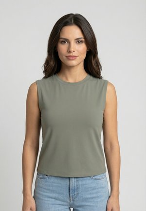 Donna con capelli lunghi e scuri che indossa una canotta verde oliva e jeans azzurri, in piedi davanti a uno sfondo chiaro uniforme.