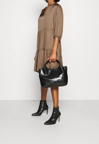 Schwarze Handtasche mit Krokodilmuster und zwei stabilen Griffen, neben einem braunen, gestuften Kleid und schwarzen Stiefeln mit glänzender Oberfläche.