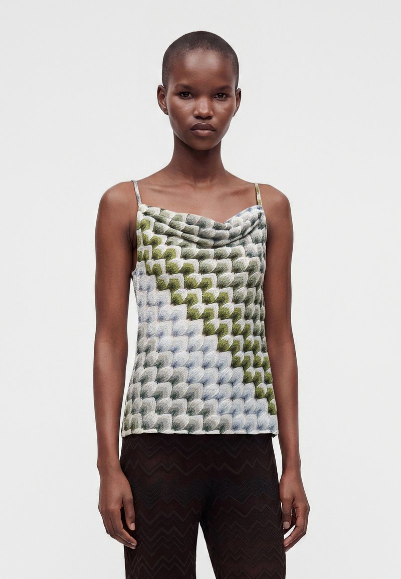 Femme portant un haut sans manches à motifs avec des écailles vertes, blanches et grises et un pantalon noir à motifs en zigzag sur un fond uni.