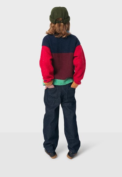 Bunt gestrickter Pullover mit roten, blauen und grünen Abschnitten, kombiniert mit gestreiften Jeanshosen und einer dunkelgrünen Mütze. Rückansicht.