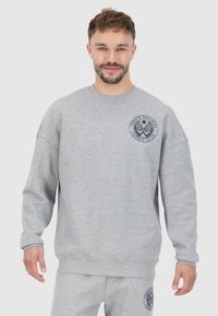 Felpa grigia realizzata in tessuto morbido con scollo rotondo. Presenta un logo blu sulla parte sinistra del petto e una vestibilità rilassata e oversize.
